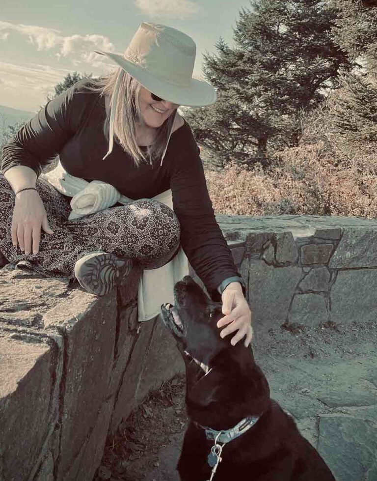 Alice pets her dog, Clyde on a mountain top.